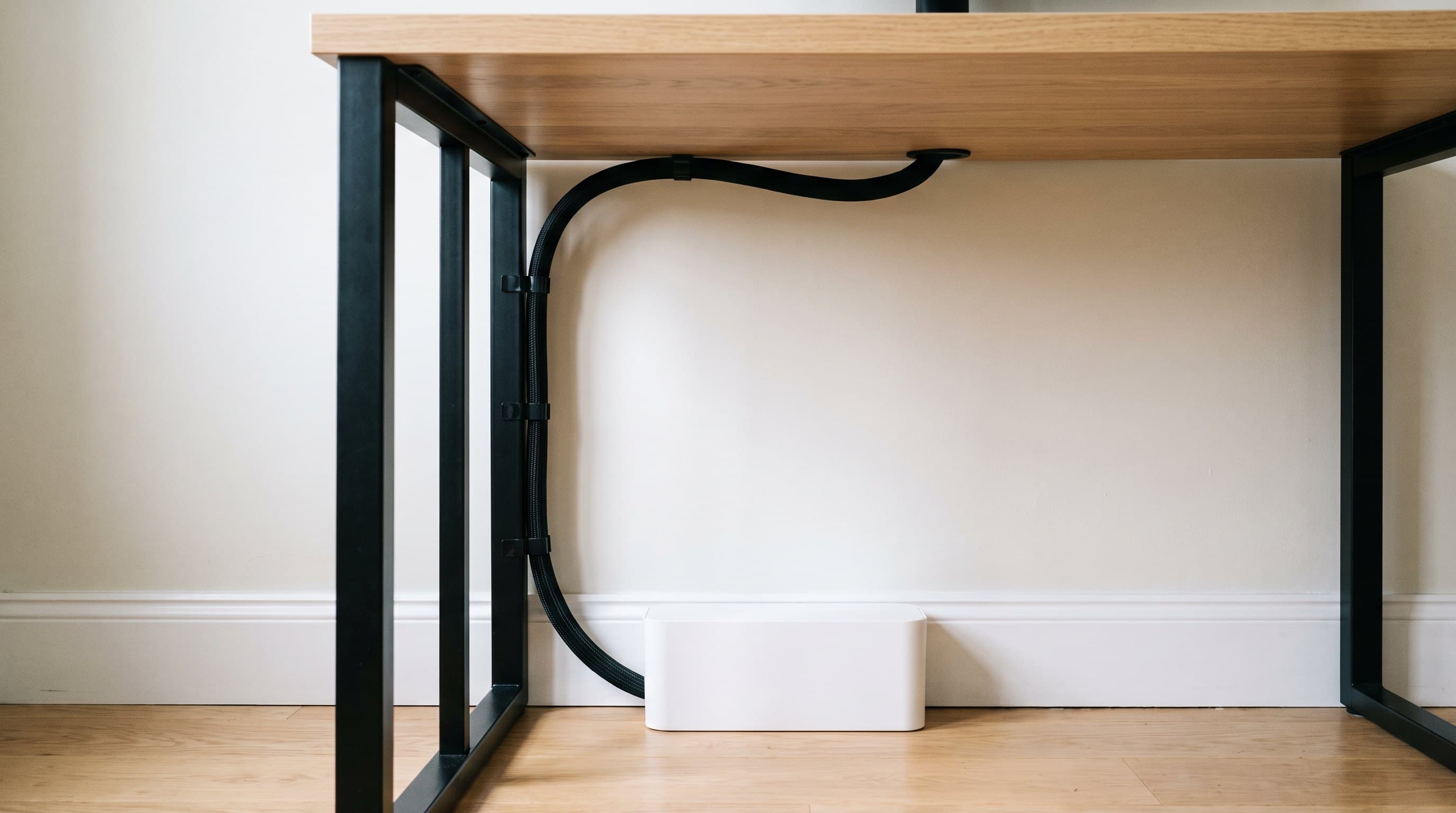 Rear view of an oak home office desk against a pale wall, a single black braided cable sleeve emerging from beneath the desktop and running down the inside of one black steel leg on three adhesive clips into a low white cable box on the floor, with no other visible cords