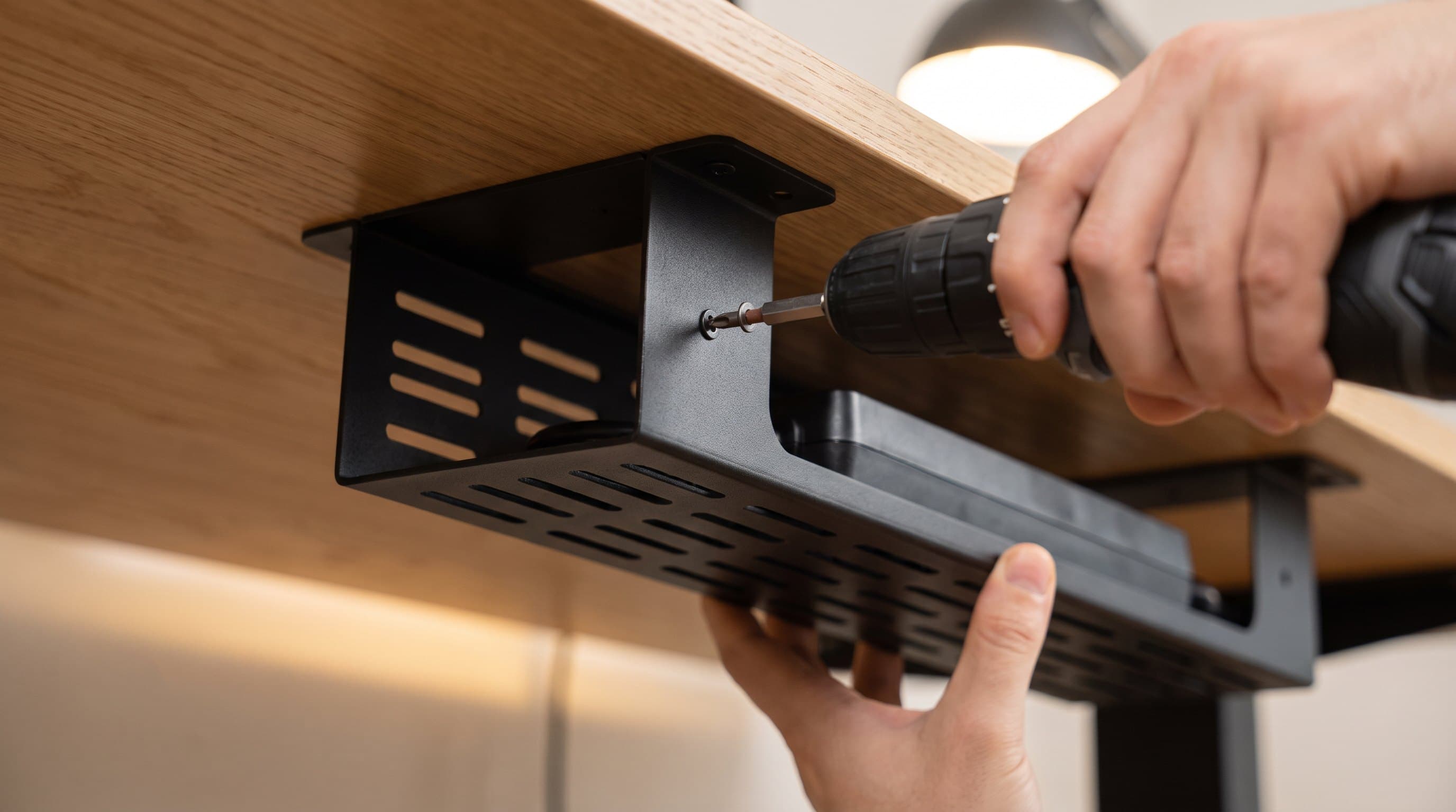 Close-up from below of a slotted black powder-coated steel cable tray held up against the underside of an oak desktop with its mounting flange flat to the surface, one hand steadying the tray from beneath while the other drives a screw home with a cordless driver, and a black power strip resting inside the tray