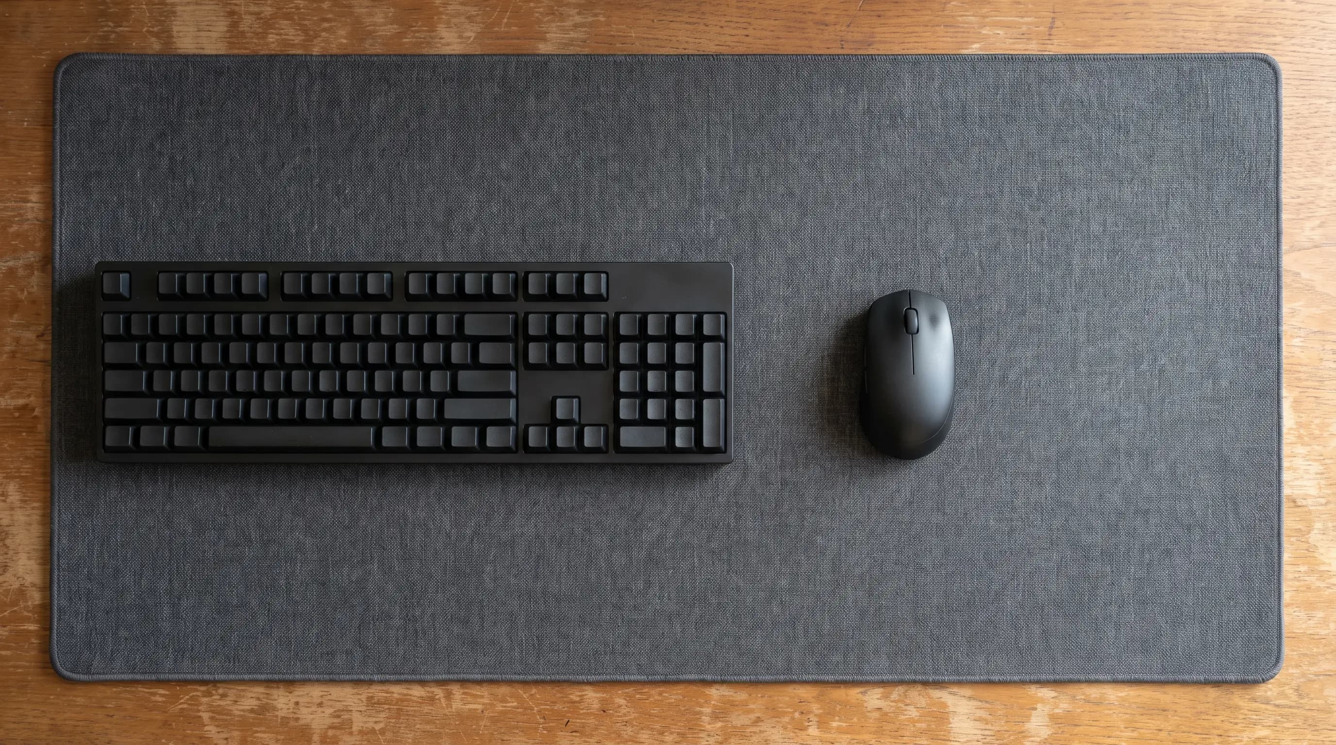 Overhead flat-lay of a large dark grey cloth desk mat on a wooden desk, a full-size mechanical keyboard with number pad occupying the left half and a black wireless mouse on the open mat to its right