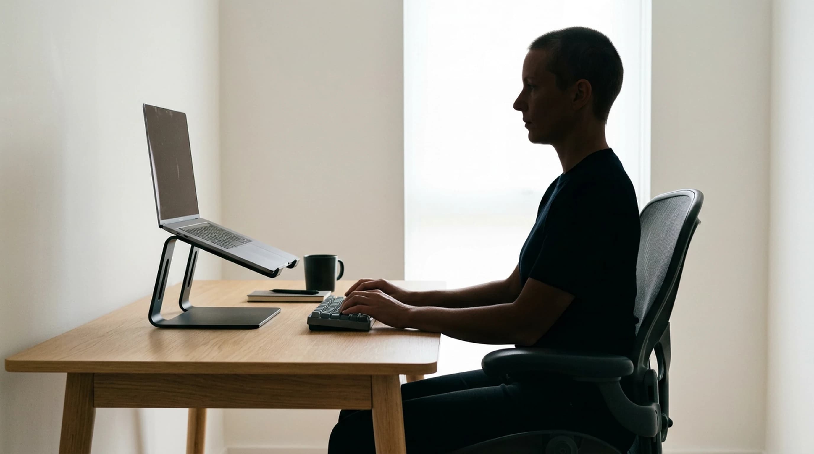 Side-on view of a person seated at a light oak desk in a bright home office, typing on an external keyboard with the laptop raised on a dark aluminum riser in front of them, their eye level visibly higher than the top of the laptop screen
