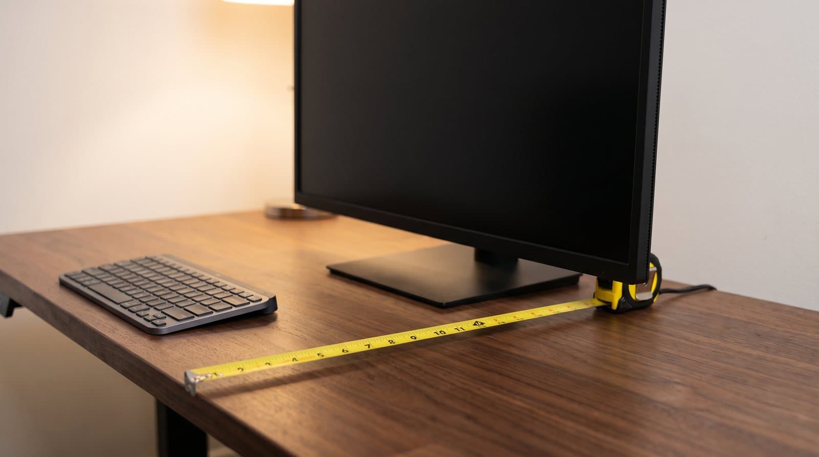 A yellow steel tape measure extended across a walnut desktop from the front edge back to the base of a black monitor, with the monitor's flat rectangular stand plate sitting on the desk beneath the screen and a compact keyboard pushed to one side