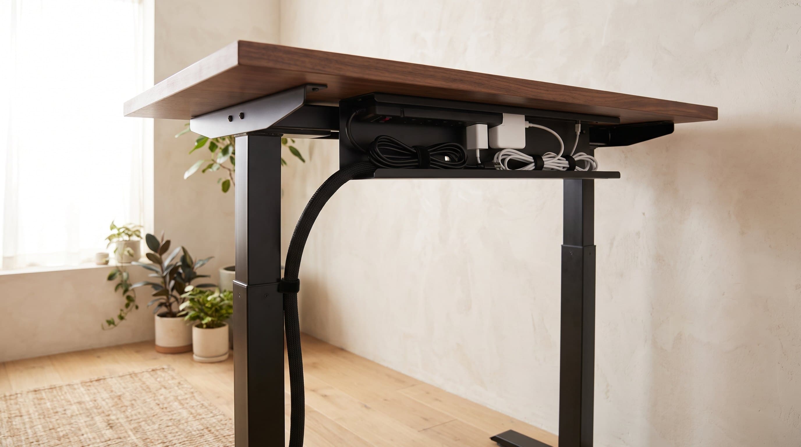 The underside of a walnut sit-stand desk in a home office, showing a black steel cable tray holding a power strip with cables gathered into a single sleeved cord dropping to the floor