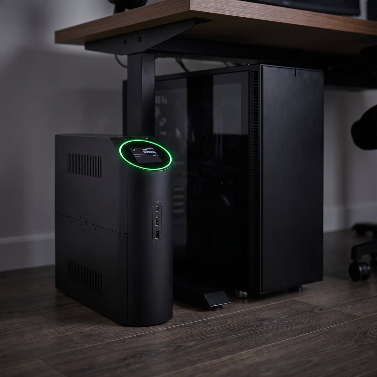 APC Back-UPS Pro Gaming BGM1500B black tower UPS with a glowing RGB ring on the front, standing beside a gaming PC under a desk