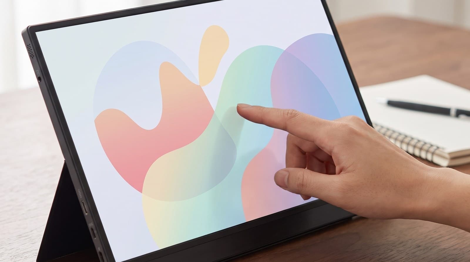 Dark 15.6-inch touchscreen portable monitor standing on a desk with a hand reaching toward the screen in a home office