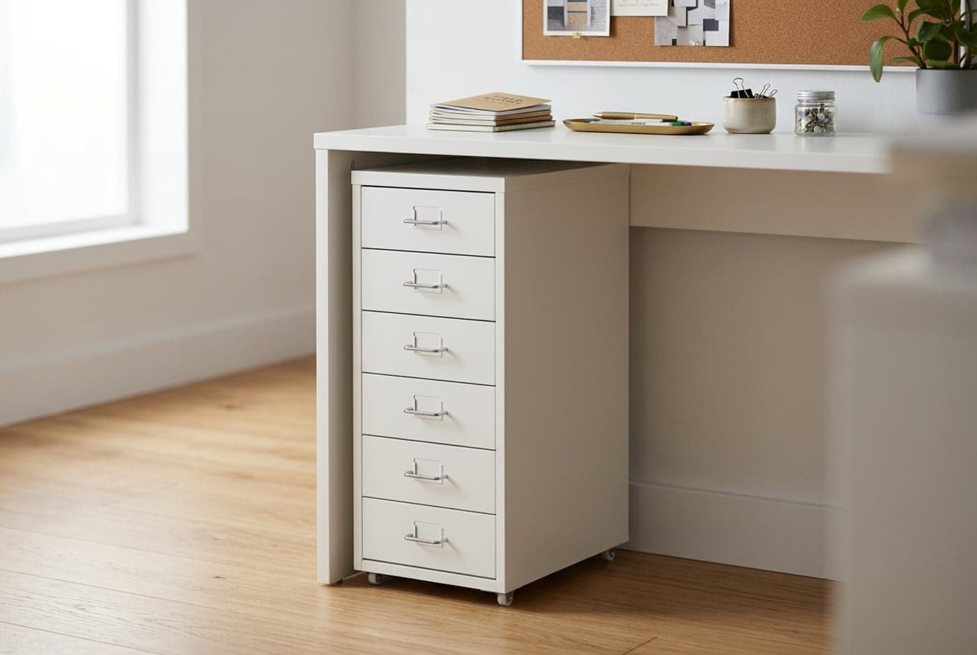 Narrow white IKEA HELMER six-drawer metal unit on casters tucked beside a desk holding stationery and small supplies