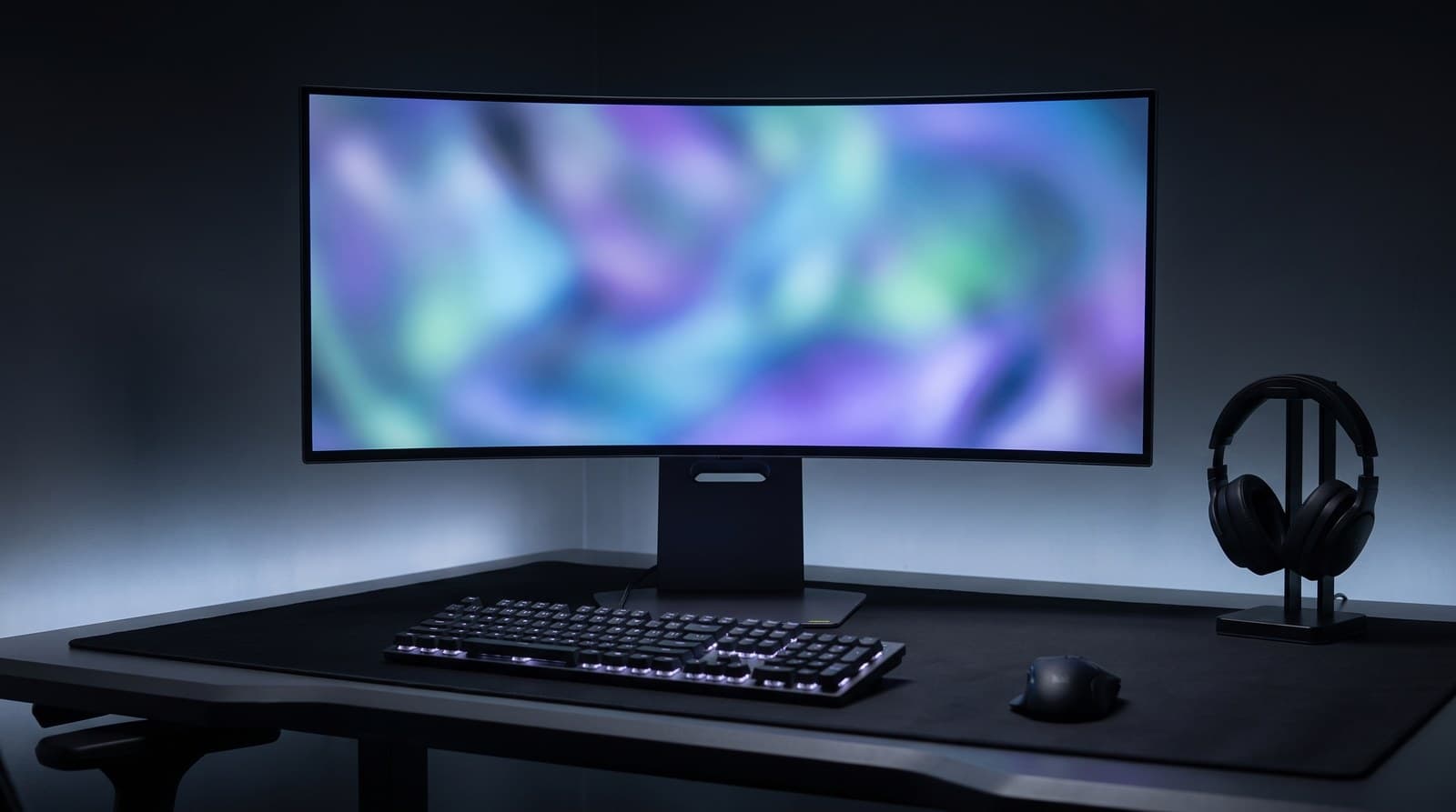 Dark grey 34-inch deeply curved OLED gaming monitor on a slotted rectangular column stand with a flat base, on a dark desk with a keyboard, mouse and headphone stand