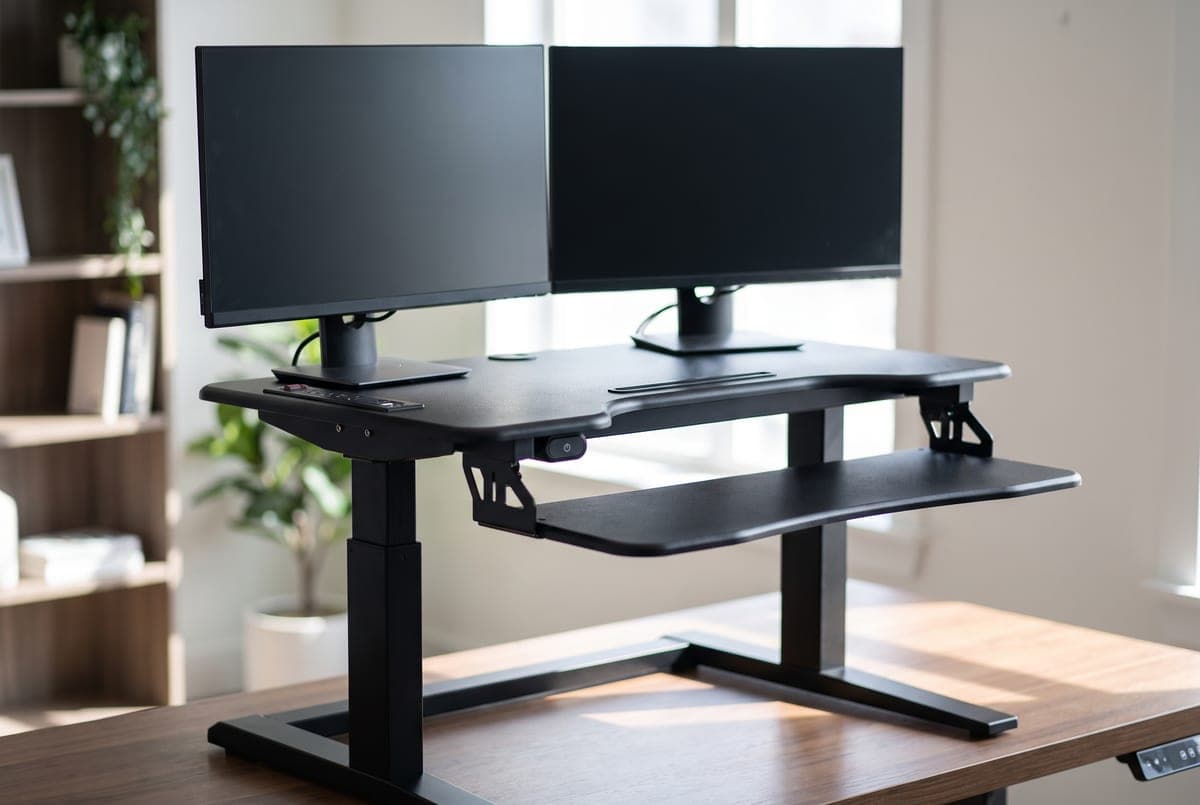 Rocelco EDR electric standing desk converter raised on a desk with a strip of power outlets along the front edge and a keyboard tray below