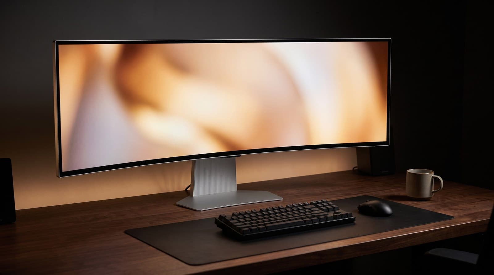 Silver 49-inch super ultrawide curved QD-OLED monitor spanning an entire desk, on a silver column stand with a wide flat base, in a dark home office with a keyboard and mouse
