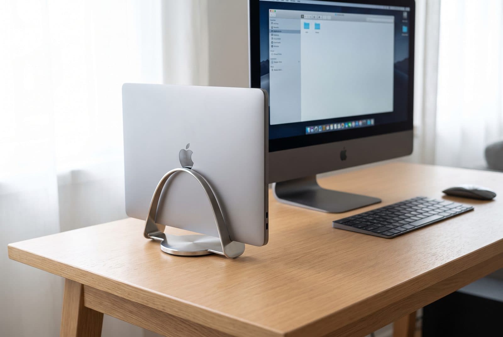 Twelve South BookArc Flex holding a closed MacBook upright by two curved steel arms on a tidy desk beside an external monitor