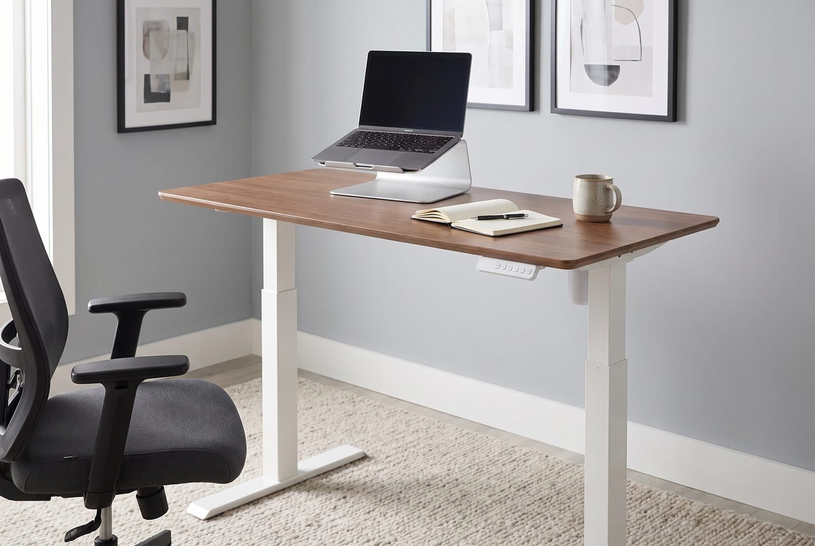 Vari Essential Electric standing desk with a hazel wood top and white T-style legs raised to standing height in a tidy home office nook with a laptop on a riser