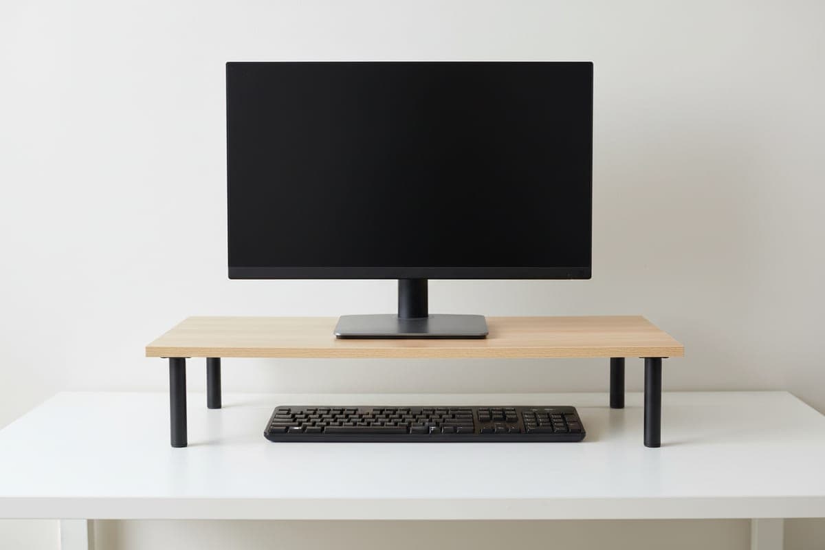 VIVO wood and steel monitor riser with a light wood top and black legs raising a monitor above a keyboard on a simple desk